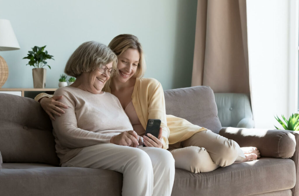 A mother and her adult daughter sitting together on a couch, smiling and looking at the daughter's phone together.