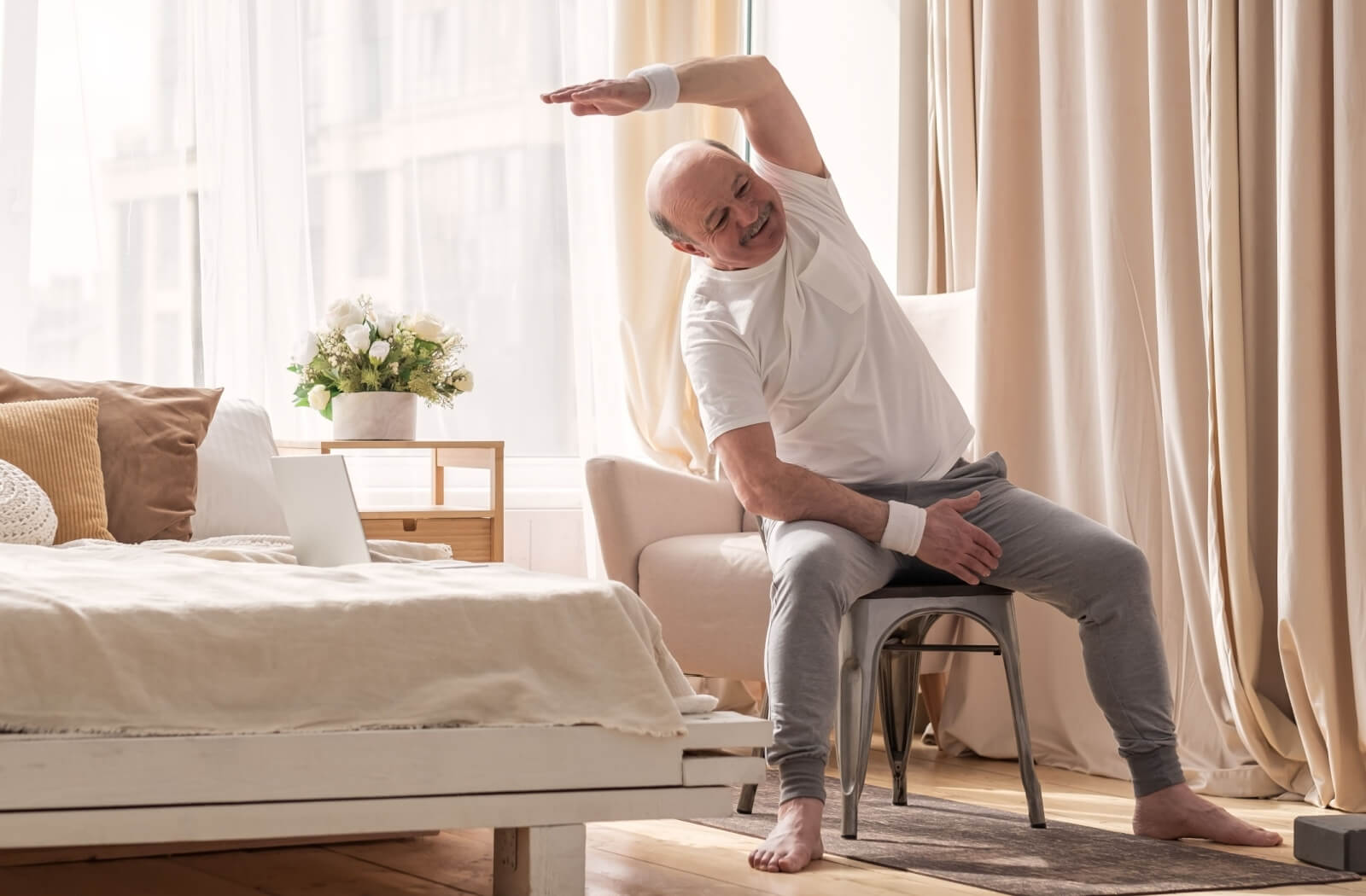 An older adult in a sunlit bedroom smiling while stretching an arm overhead during a chair core exercise.