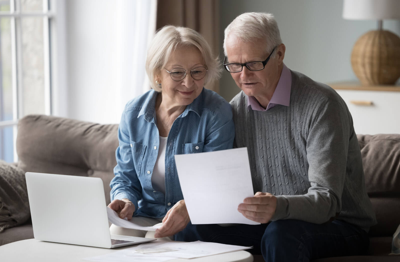 A retired couple on their couch with a laptop and paperwork, working together to determine if they meet the eligibility requirements for assisted living.