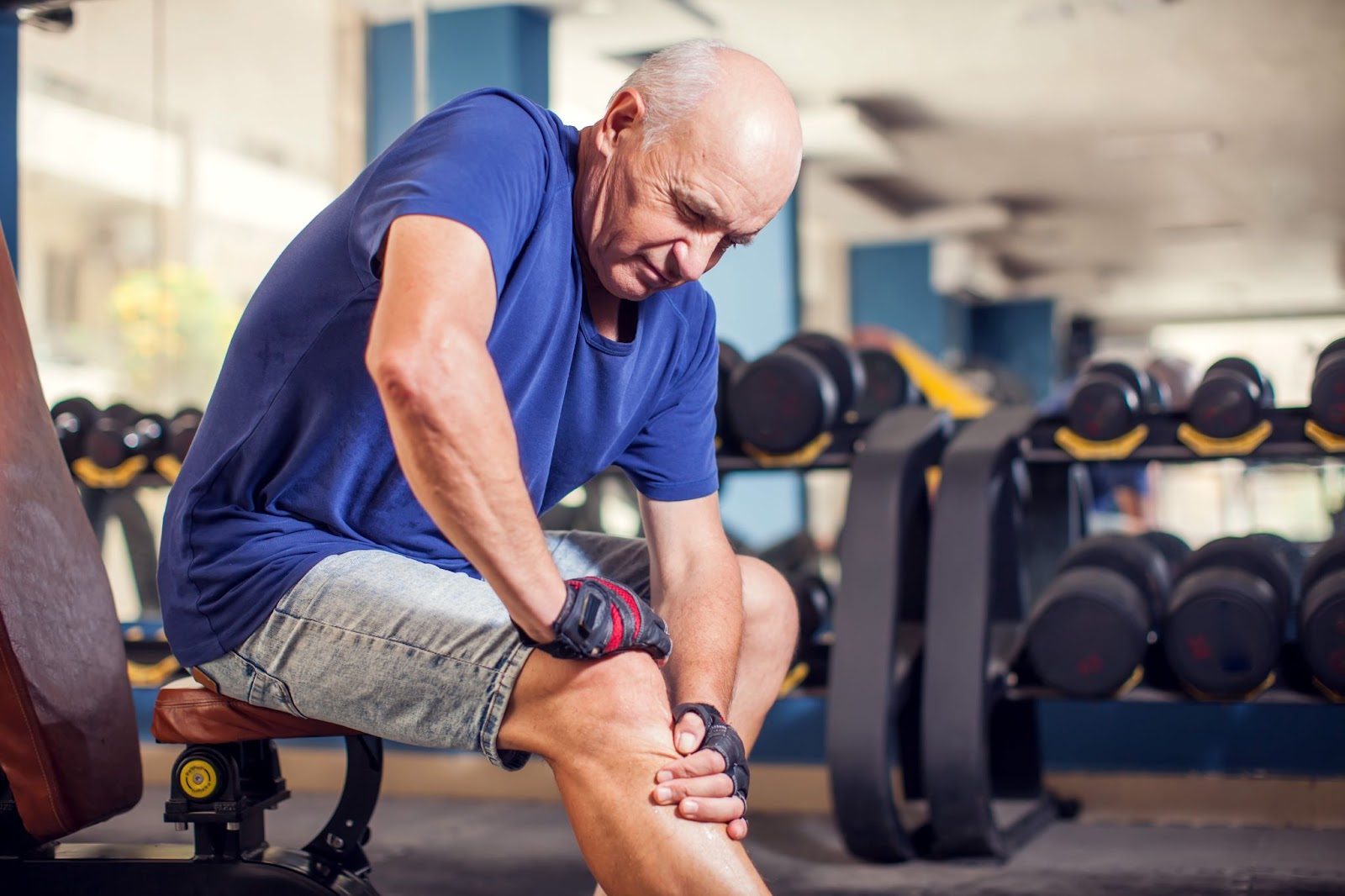 An older adult expressing pain while sitting on a bench in the gym holding his right knee.