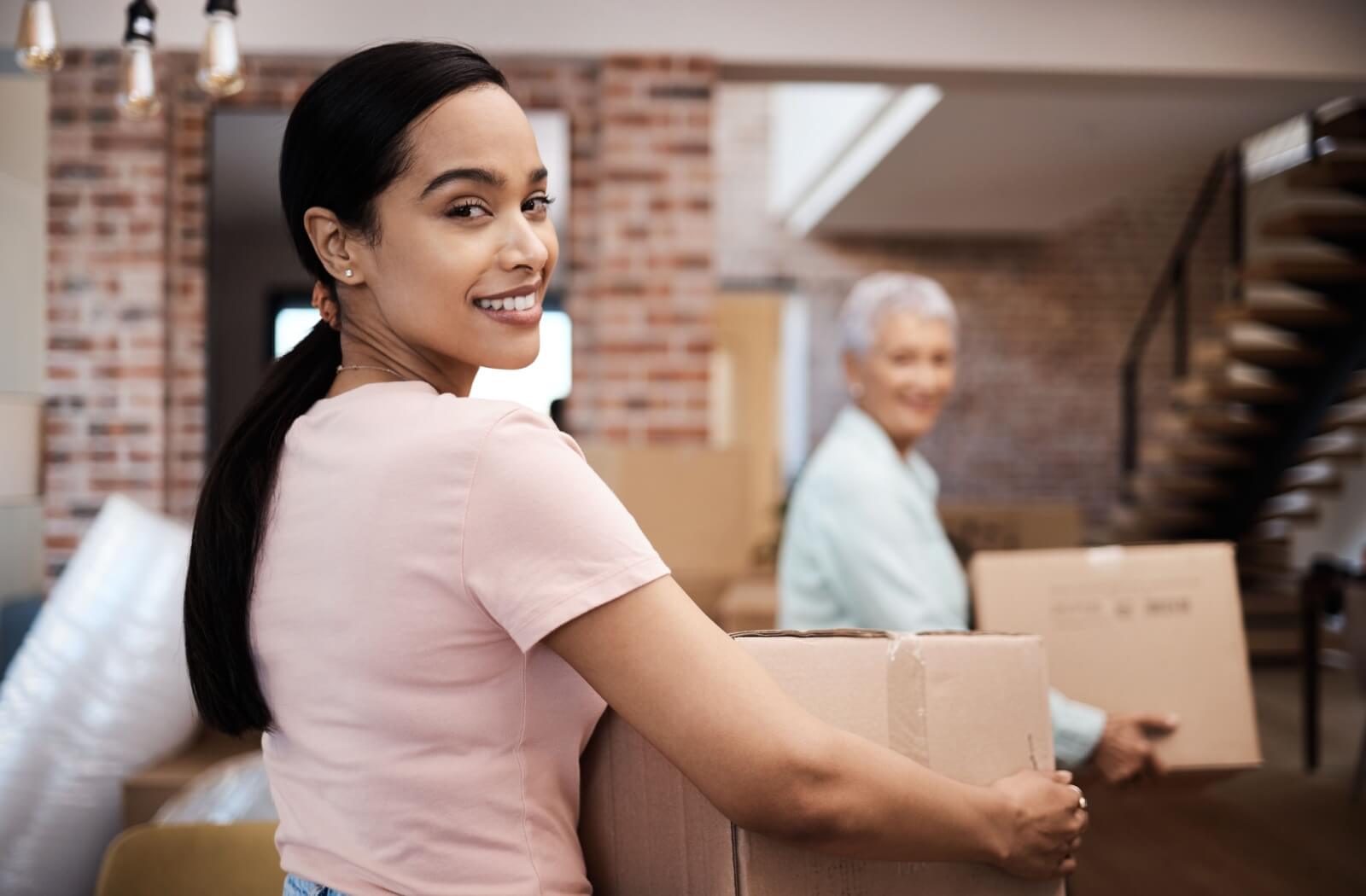 An adult daughter smiling and helping her older mother carry moving boxes