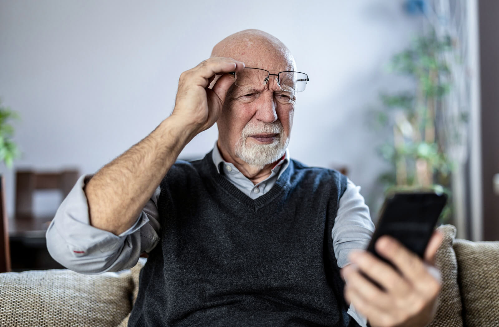 An older adult lifting their glasses in confusion and looking at their cell phone in confusion due to a senior scam call.