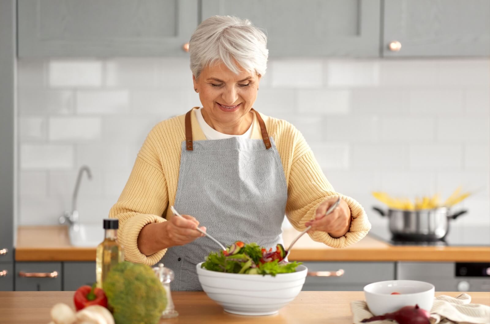 A smiling older adult wearing a kitchen apron mixing salad in a white bowl.