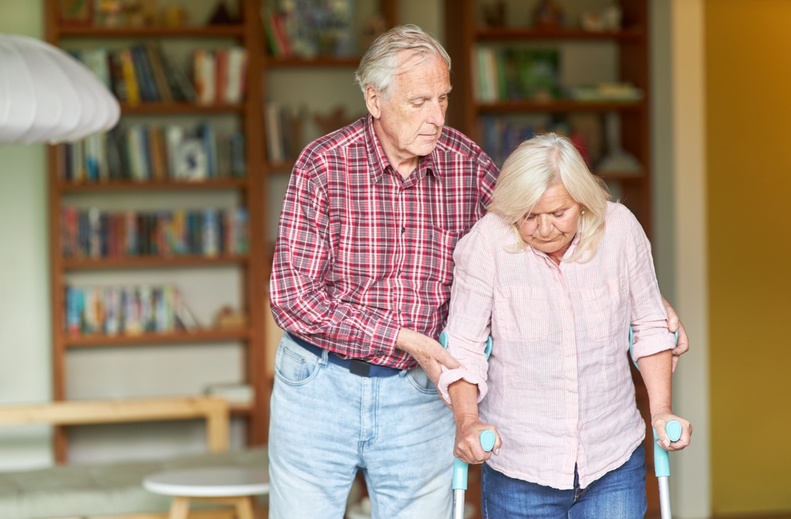 2 senior spouses walking together while one supports the other as they use forearm crutches to walk.