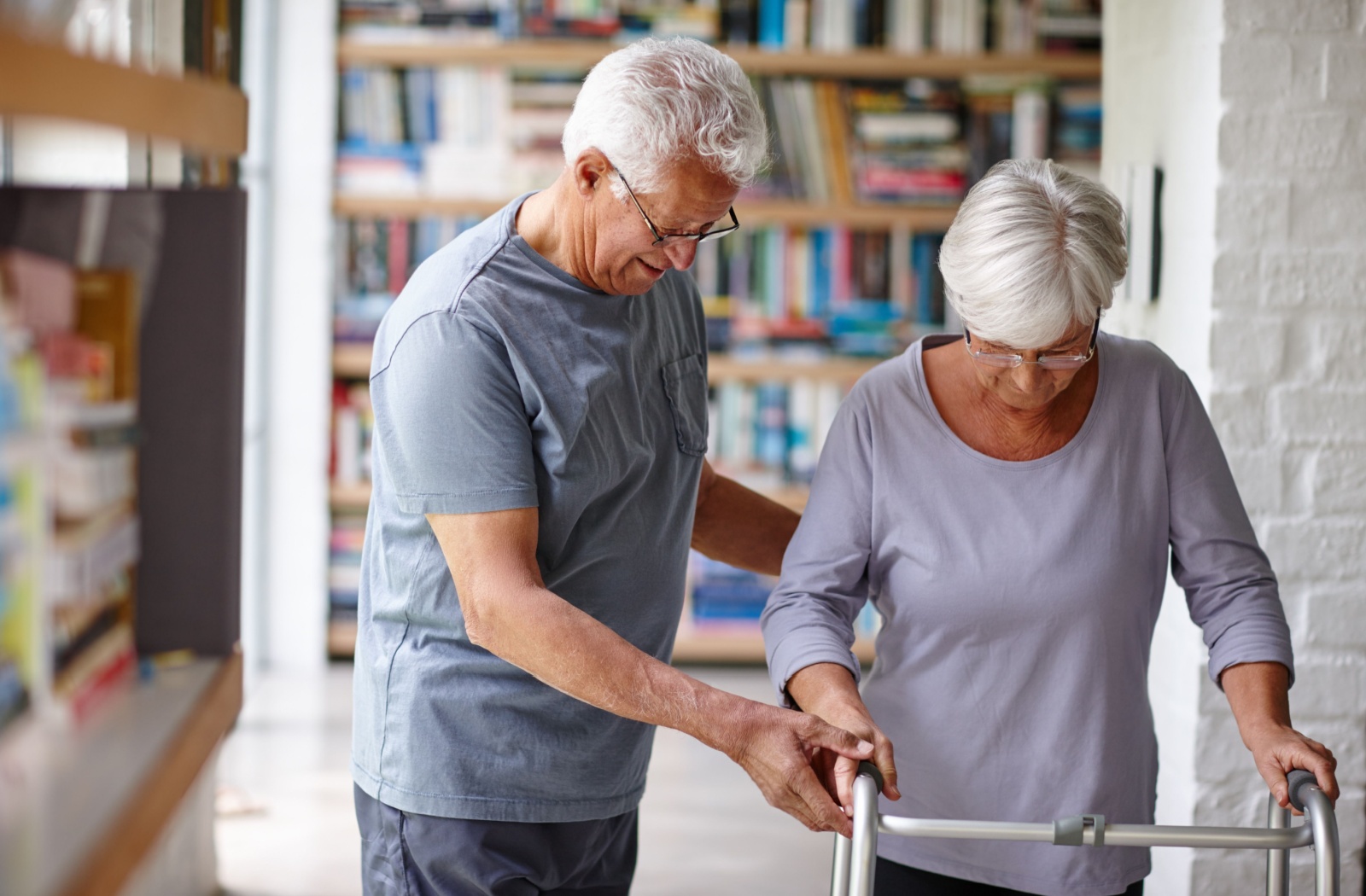 Two older adult spouses walking together while one helps the other use an assistive walker.