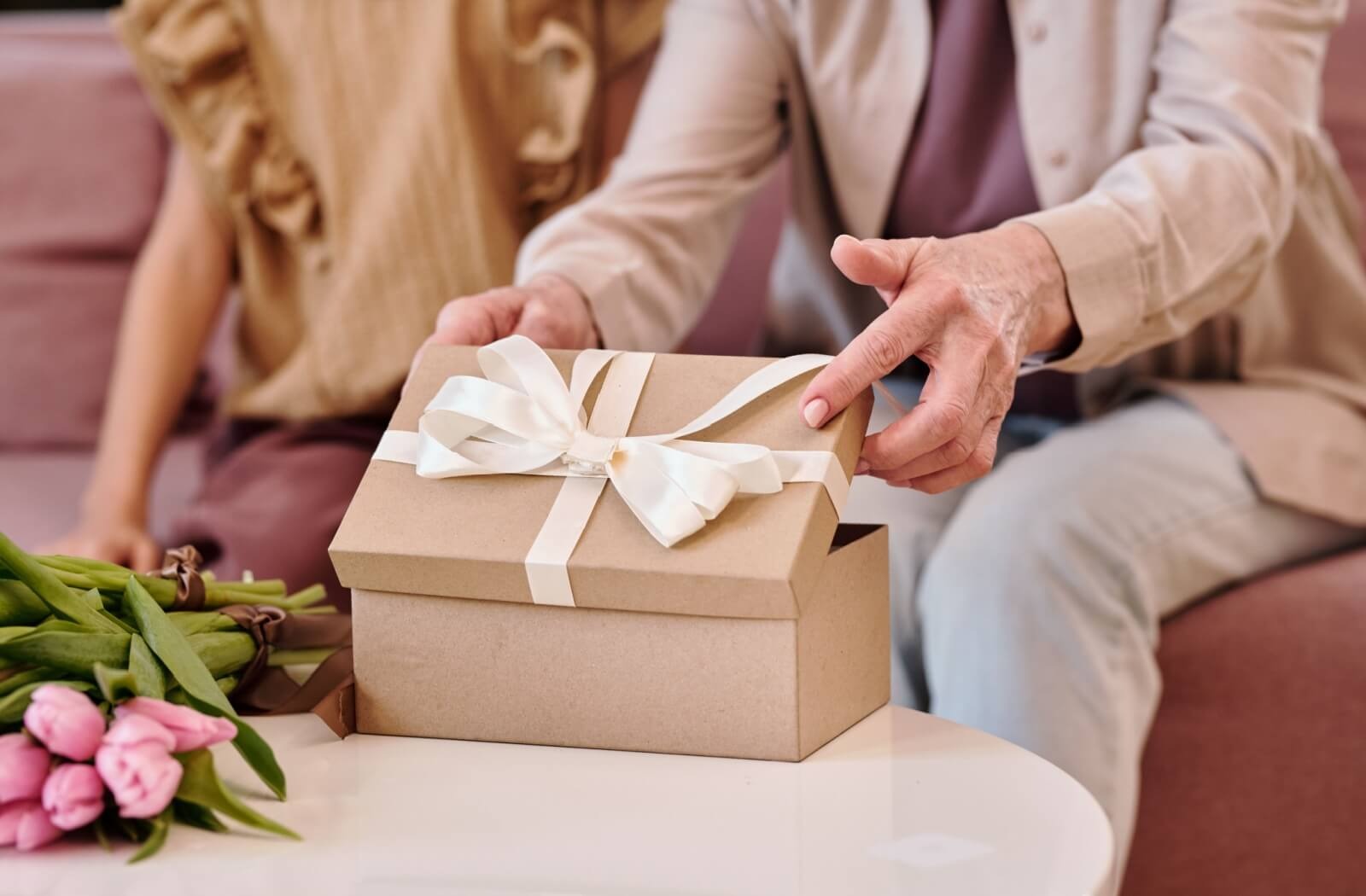 An older adult sits beside their grandchild and opens a carefully-wrapped box with a bow
