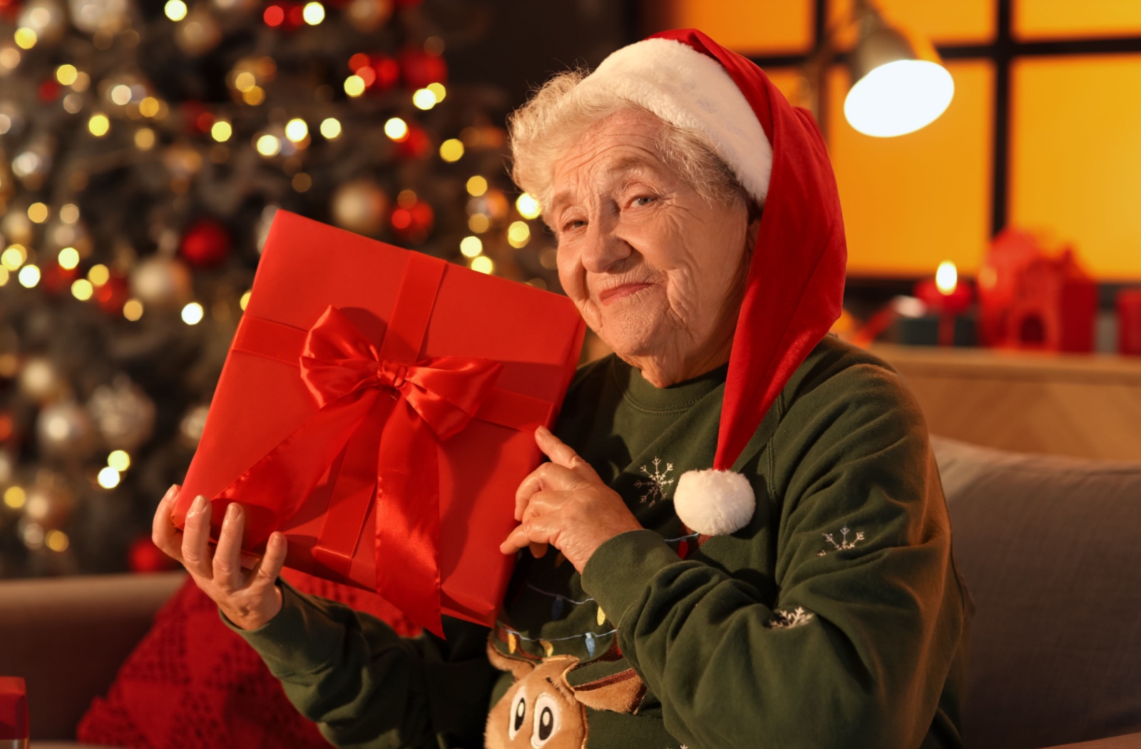 An older adult wearing a Santa hat smiles and holds up a red gift wrapped in a red bow in front of a Christmas tree